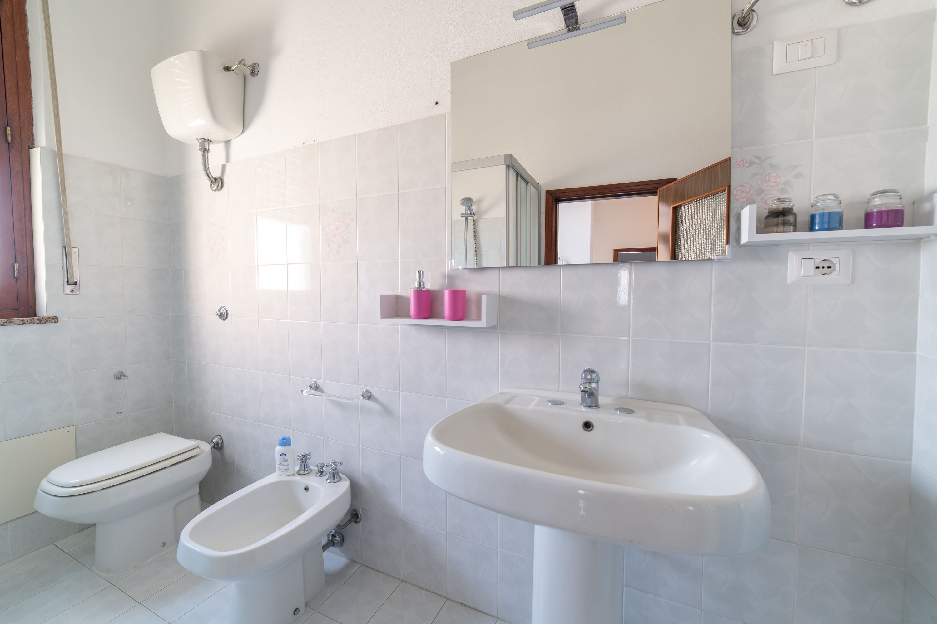 A bathroom with a toilet, bidet, sink, mirror, and shelves with toiletries.