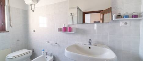 A bathroom with a toilet, bidet, sink, mirror, and shelves with toiletries.