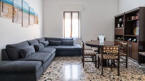 Een woonkamer met een hoekbank, een eettafel met stoelen, een boekenkast en een raam met gordijnen.