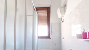Een badkamer met een douchecabine van glas, een toilet, een bad en een raam met zonwering.