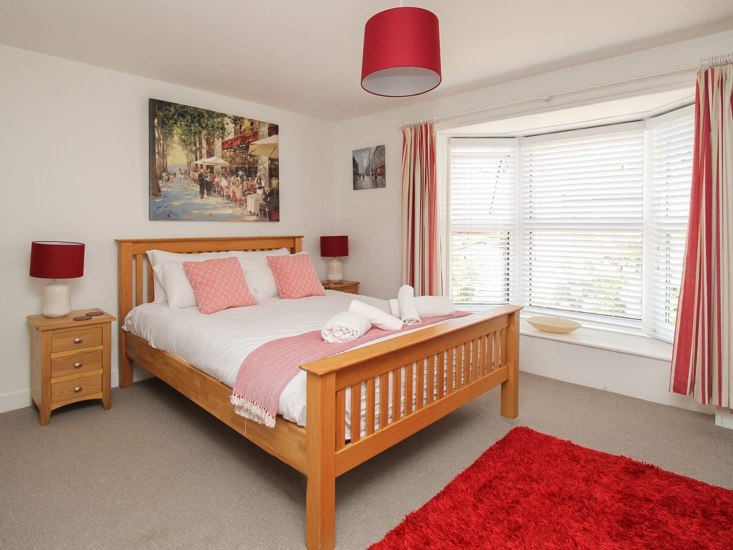 A bedroom with a wooden bed, two bedside tables, a red rug, and a large window with curtains.