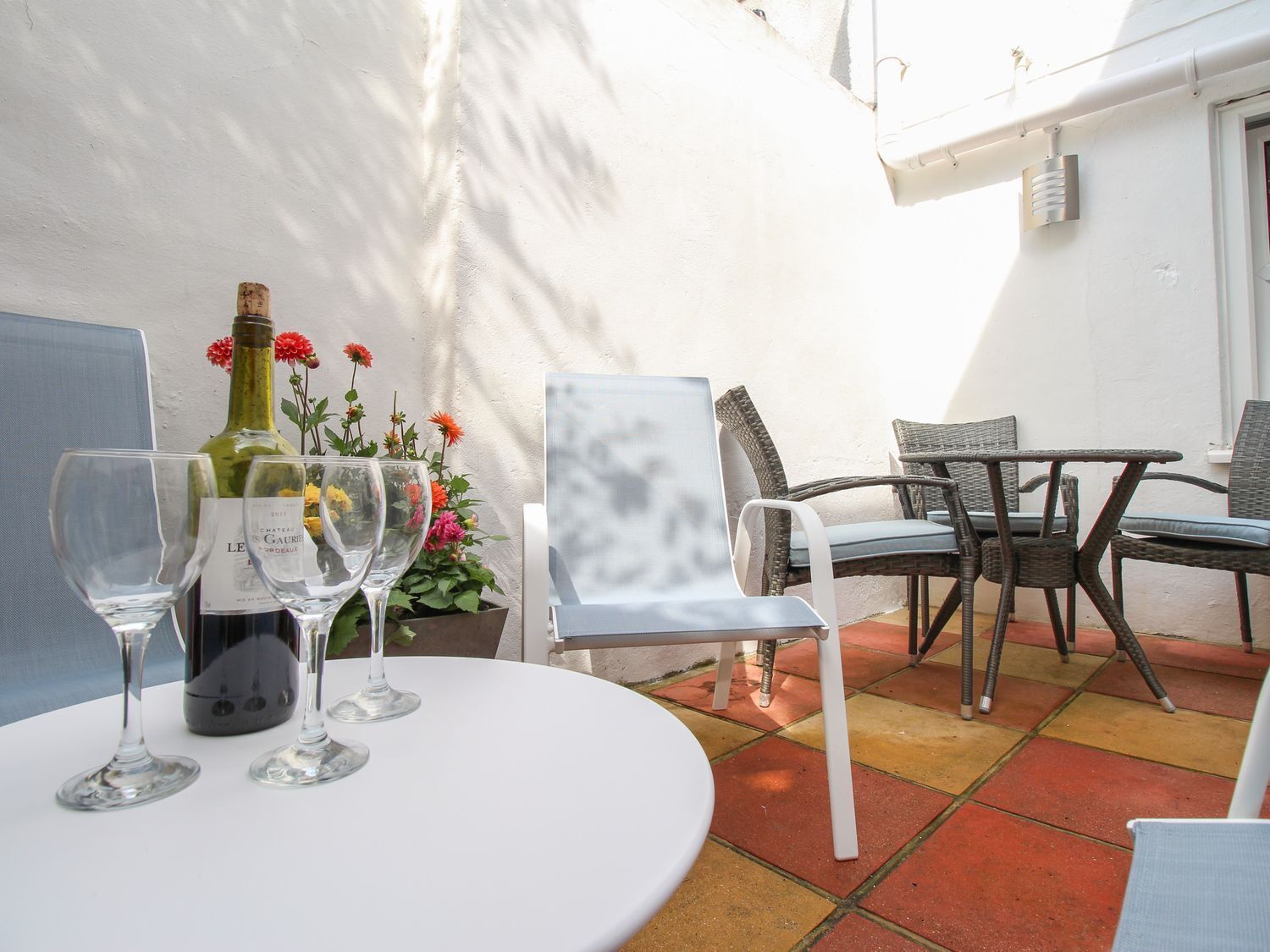 A patio with a round table, two chairs, a bottle of wine, and a potted plant with flowers.