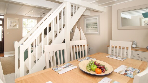 A dining area with a wooden table, white chairs, a plate of fruit, and a staircase with white railings. - Little Gull Cottage (Weymouth)