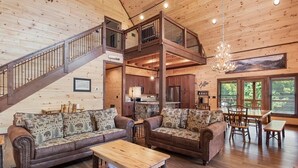 A spacious cabin interior with a high ceiling, wooden floors, a staircase, a dining area, and a kitchen. - Gorgeous 4BD w/ VIEWS! Theater +Firepit +Game Room (Sevierville)