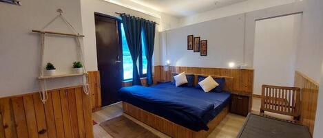 A bedroom with a bed, wooden headboard, bedside tables, a small wooden chair, a hanging plant, and a window with curtains.