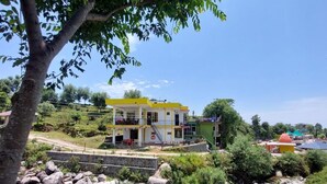 A two-story building with a yellow and white facade, situated on a hillside with a rocky foreground and a small stream. - The Hostel Stories Bir - Landing Site (Baijnath)