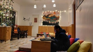 A cozy room with a wooden desk, a laptop, and a colorful couch. There is a wall with a travel-themed tapestry and a sign that says "Travel with us." - The Hostel Stories Bir - Landing Site (Baijnath)