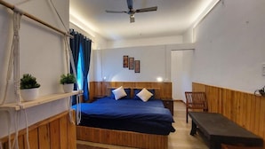 A bedroom with a bed, bedside tables, a ceiling fan, a bench, and a window with curtains. - The Hostel Stories Bir - Landing Site (Baijnath)