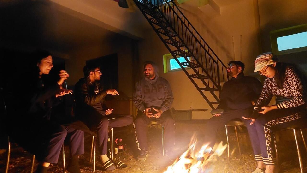 A group of people sitting around a campfire in a dimly lit room with a staircase in the background.