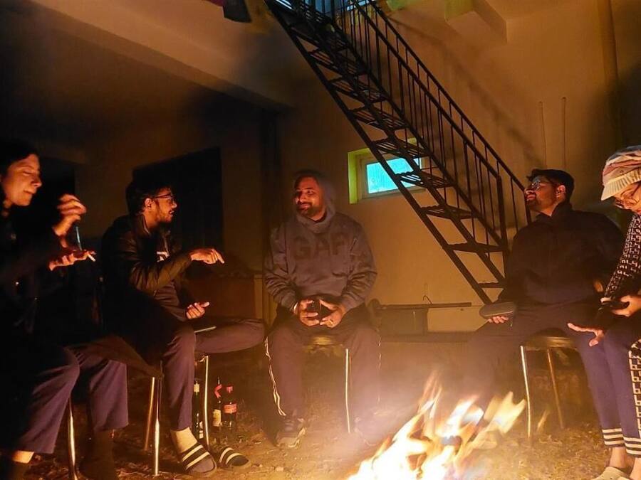 A group of people sitting around a campfire in a dimly lit room with a staircase in the background.