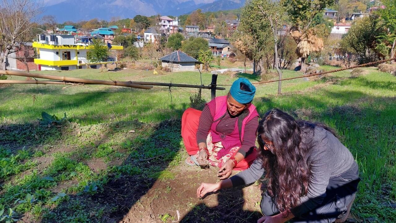 Two people sitting on the ground in a rural setting with houses and mountains in the background.