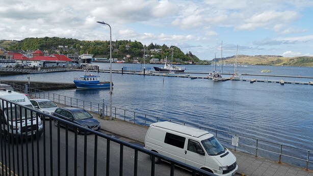 A coastal marina with boats docked, a white van parked on the side, and a clear view of the water and hills in the background. - Bay View Esplanade Apartment Overlooking Harbour (Oban)