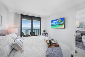 A hotel room with a bed, a view of the sea, a painting of a beach, and a tray with a bottle and glasses. - Breath-Taking Beachfront Condo | Pool/Spa/Views (Marco Island)
