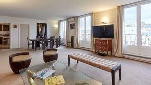 Amplia sala de estar con una mesa de centro de vidrio, dos sillones, un banco, un televisor y una zona de comedor con una mesa y sillas.