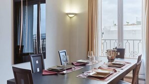 Un comedor con una mesa larga preparada para cuatro personas, una ventana grande con cortinas, y una lámpara fijada a la pared.