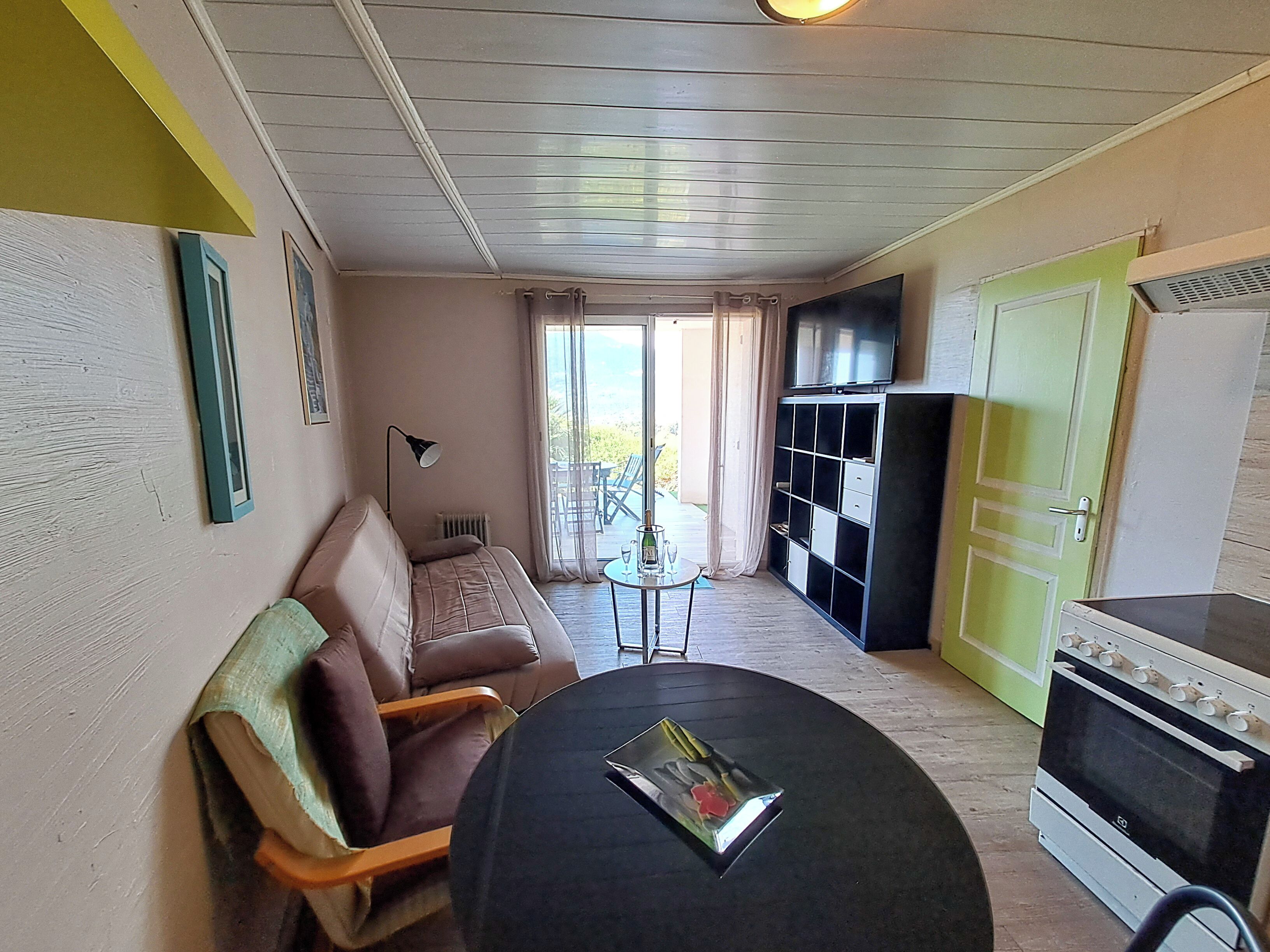 A compact living space with a sofa, a small table, a kitchenette, and a sliding glass door leading to an outdoor area.