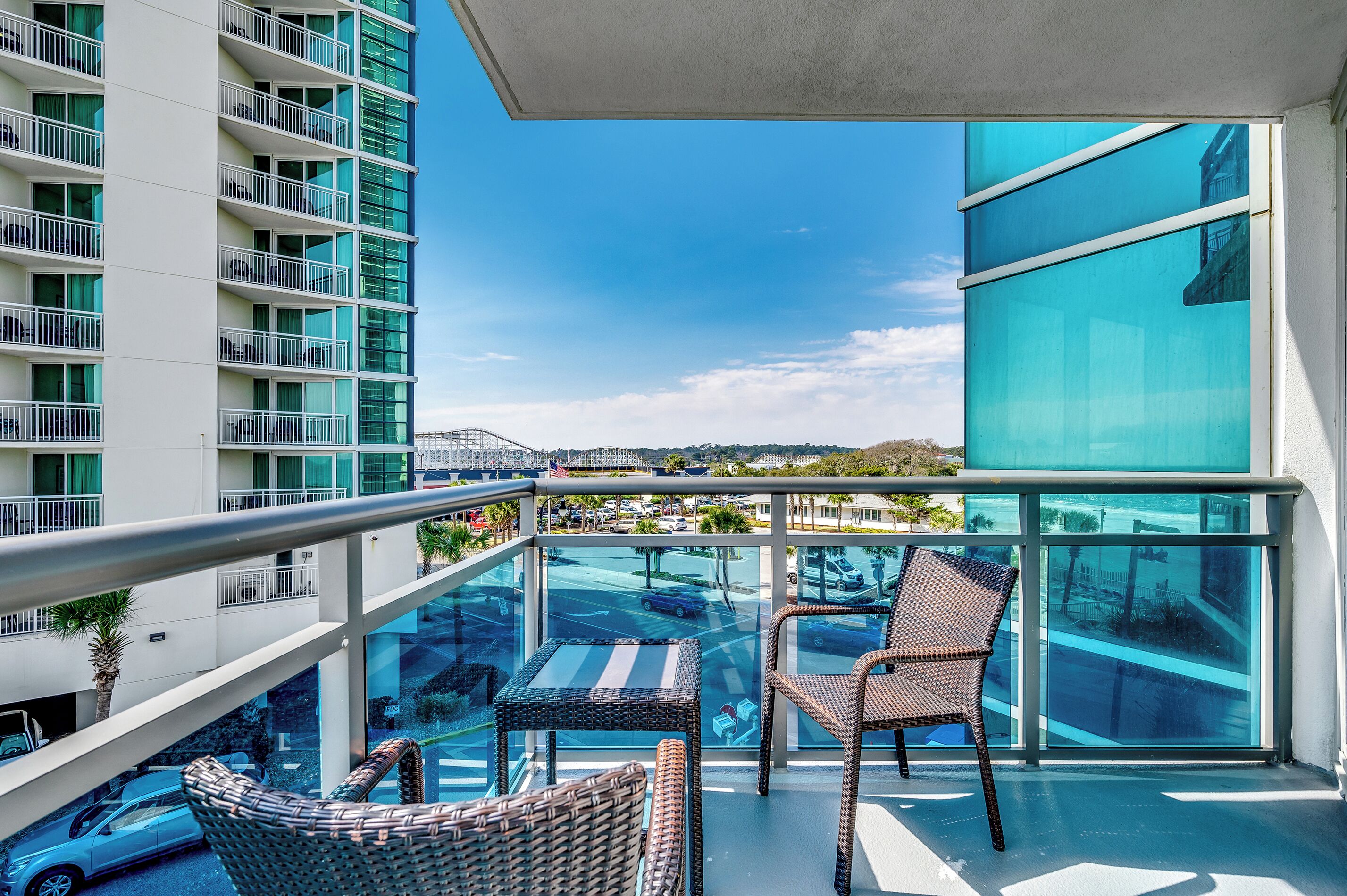 A balcony with wicker chairs, a glass railing, and a view of a parking lot and buildings.