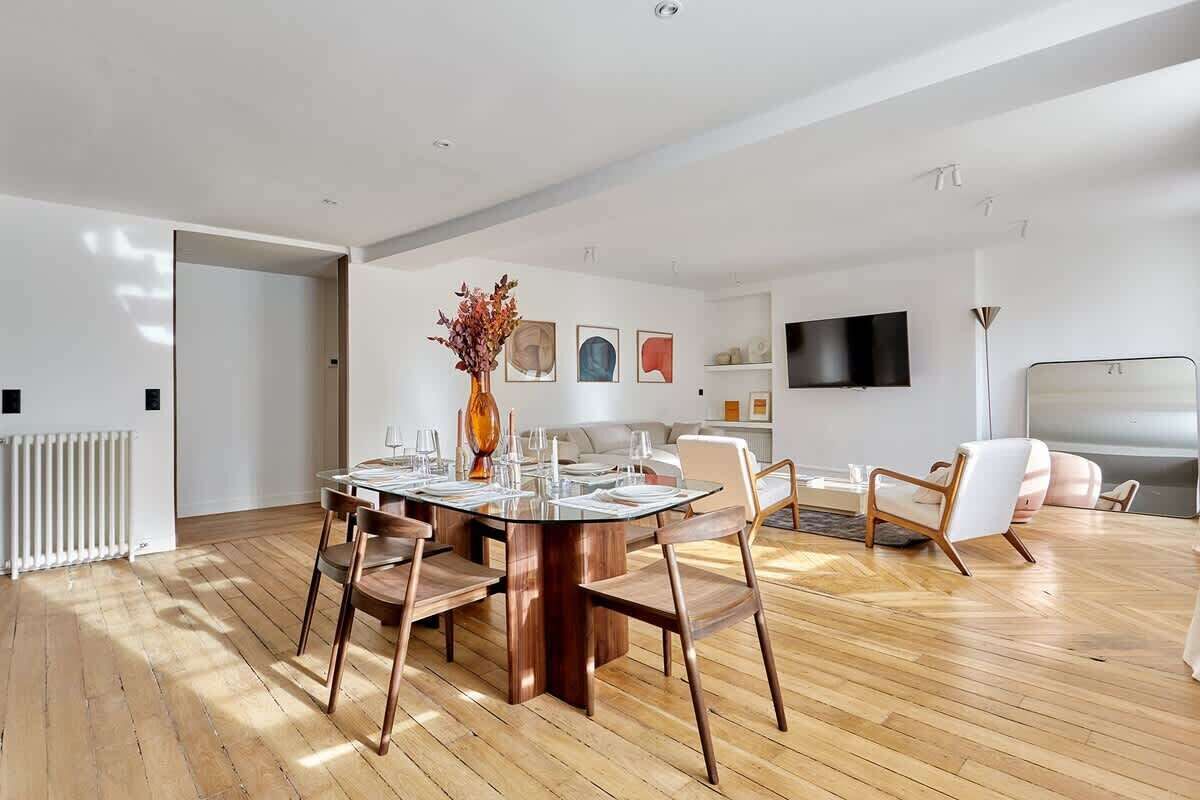 A spacious living area with a dining table set for four, surrounded by modern chairs. A flat-screen TV is mounted on the wall, and a sofa is visible in the background.