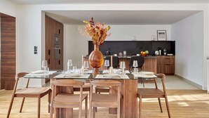 A modern dining area with a glass table, wooden chairs, and a vase with dried flowers. A kitchen with dark cabinets and a black countertop is visible in the background. - Luxurious apartment Louvre Museum-Palais Royal (Paris)