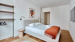 A modern bedroom with a bed, a bedside table, a wall-mounted shelf, and a door leading to another room. - Luxurious apartment Louvre Museum-Palais Royal (Paris)