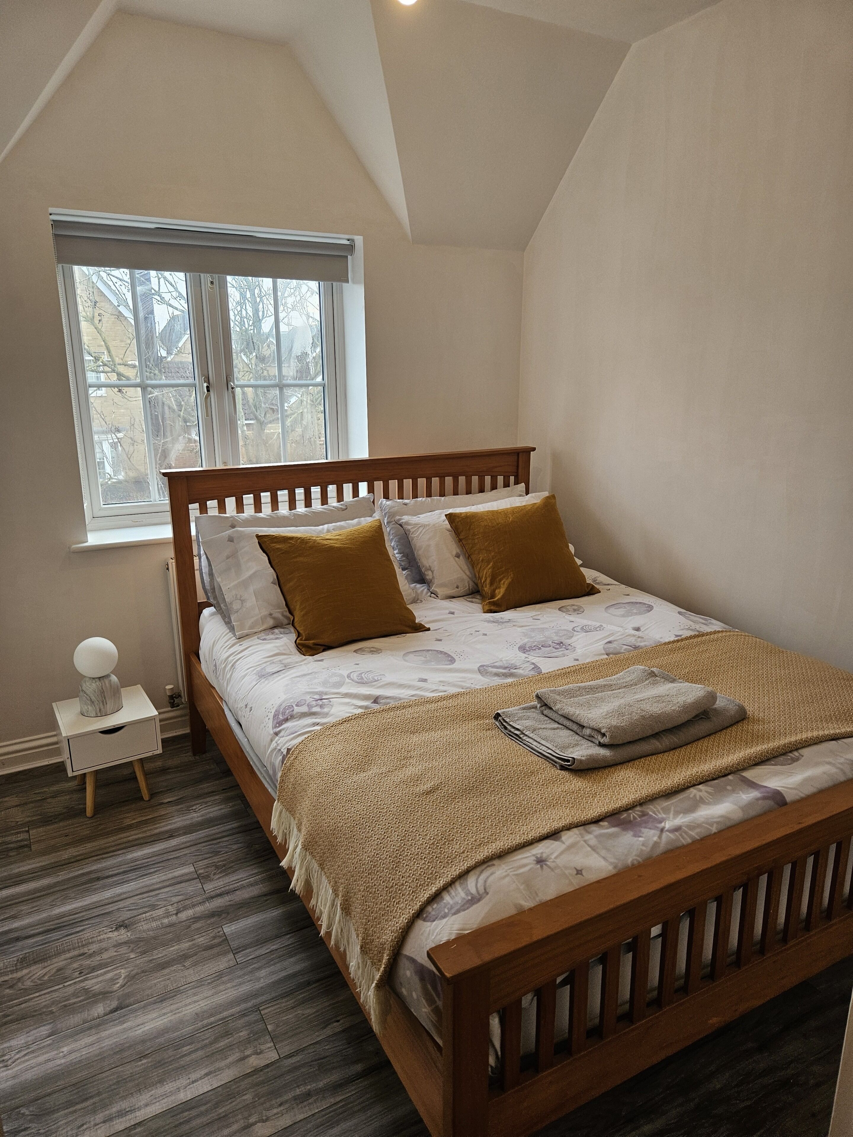 A bedroom with a wooden bed, a nightstand, a window with blinds, and a view of the outside.