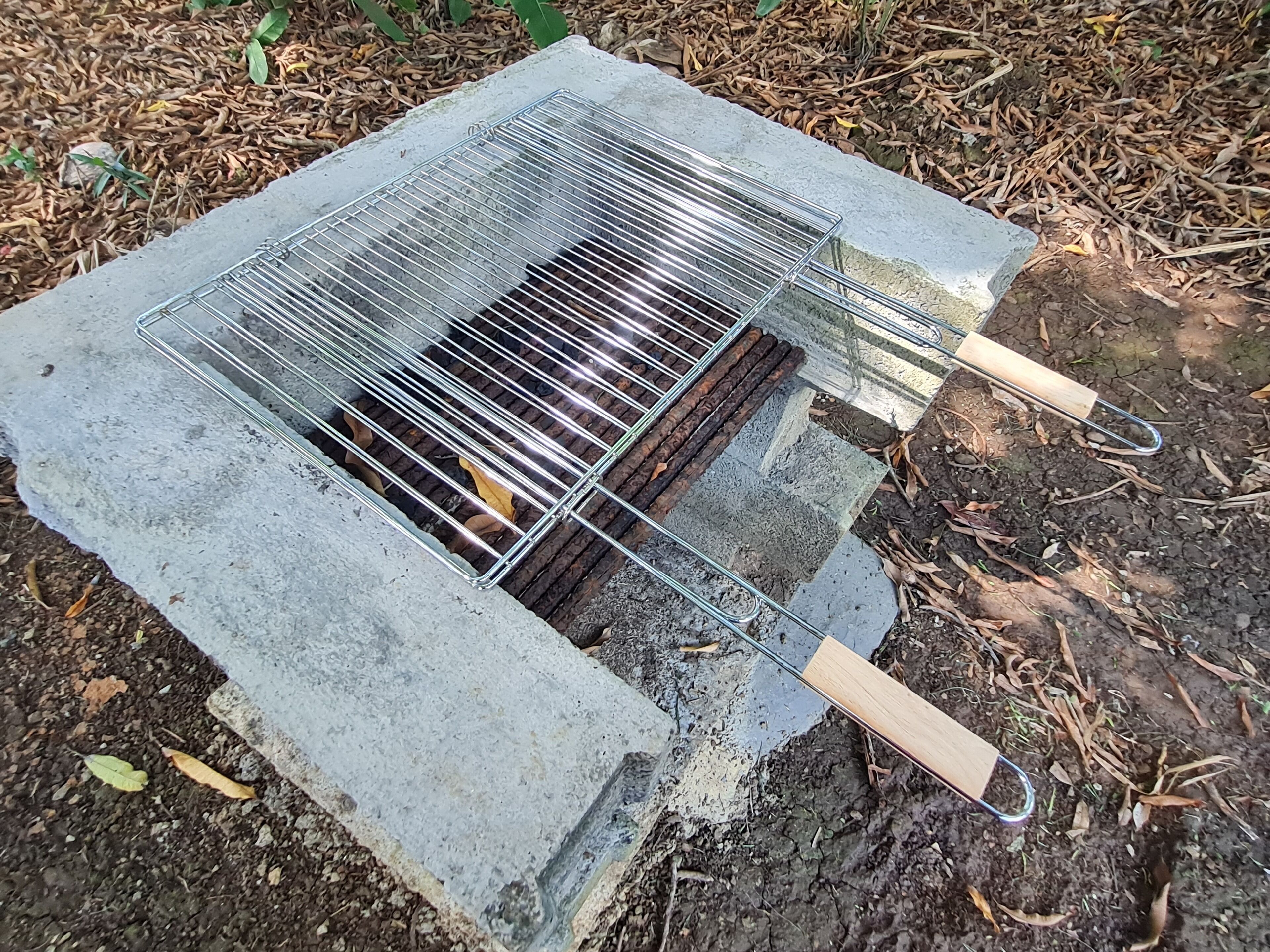 A portable grill with a metal grate and wooden handles, placed on a concrete base outdoors.