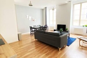 Un soggiorno moderno con un divano scuro, pavimento in legno, zona pranzo con sedie e tavolo, televisione e ampie finestre.