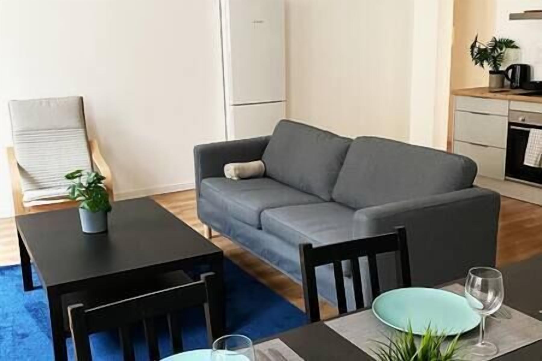 A modern living room with a grey sofa, a small dining table with two chairs, and a kitchen area in the background.
