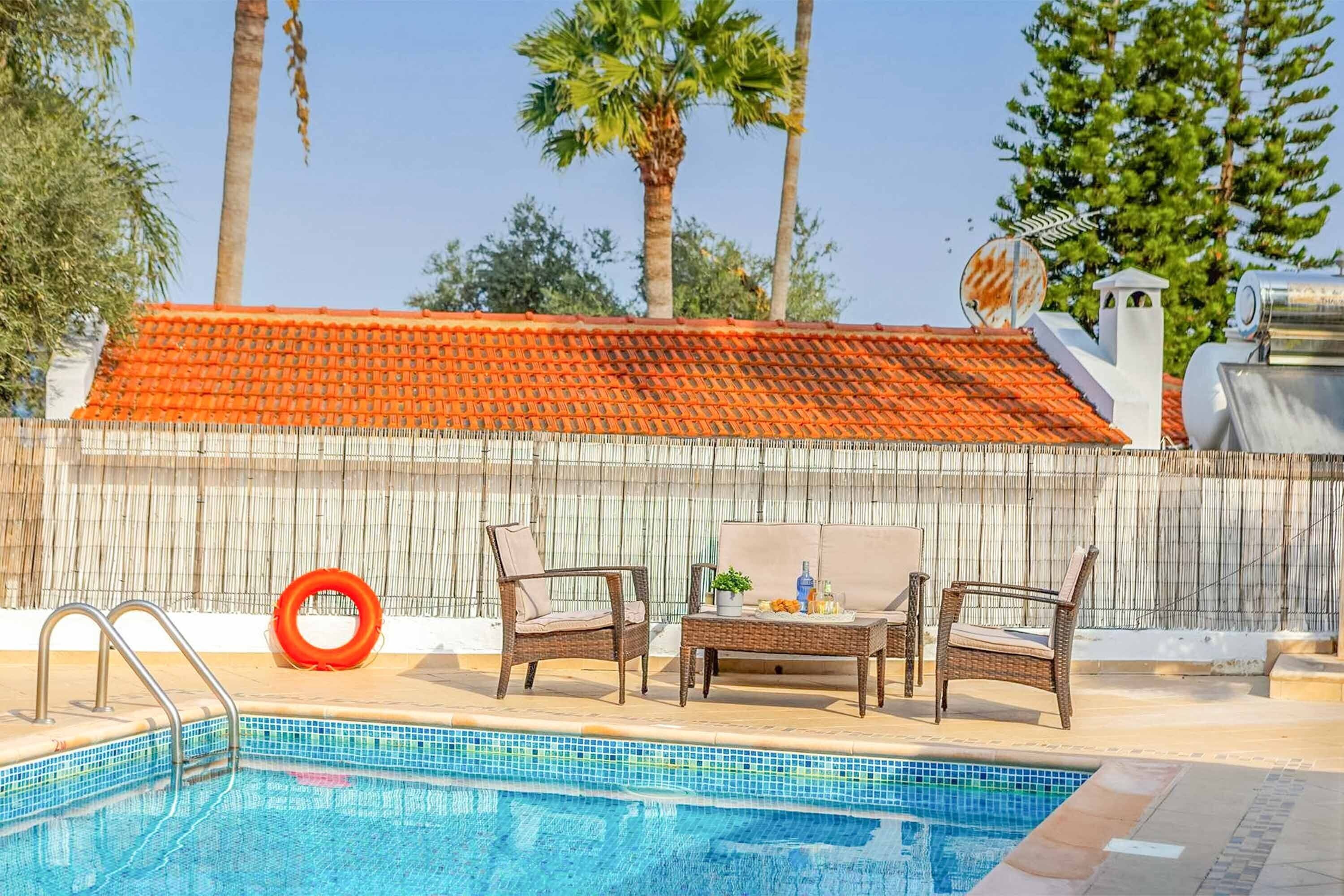 Une piscine entourée d’une clôture en bambou et de palmiers, avec des chaises longues et une table à proximité.