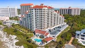 A high-rise hotel with multiple balconies, a swimming pool, and a parking area. - Beach Daze - Beach Manor 113 | Ground Floor Condo | Steps to Beach | Pool, Sauna & More! (Miramar Beach)