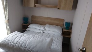 A neatly made bed with white linens, a wooden headboard, and two bedside tables with teal lamps. - 208 Unity Beach 6 Berth enc Garden no Pets (Burnham-on-Sea)