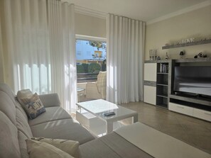 A modern living room with a sofa, a coffee table, and a TV. There is a balcony with outdoor furniture and a view of buildings.
