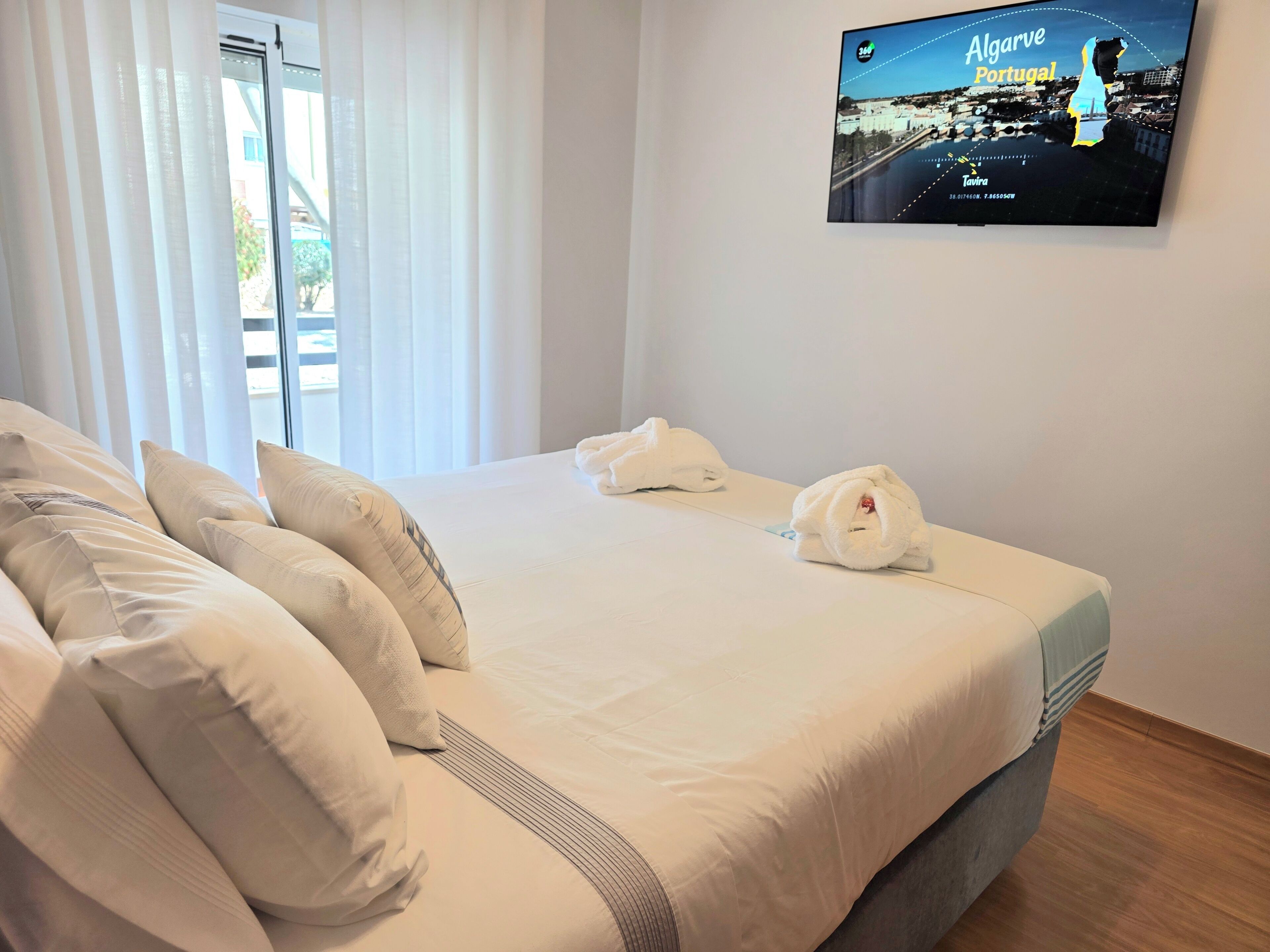 A hotel room with a bed, pillows, a TV screen displaying a cityscape, and a window with curtains.