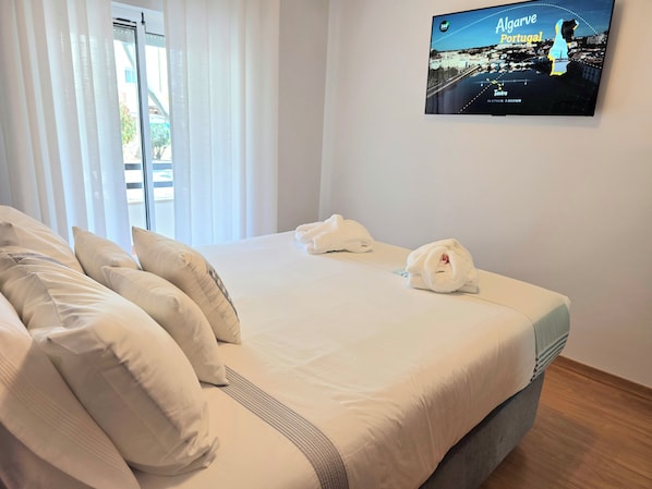 A hotel room with a bed, pillows, a TV screen displaying a cityscape, and a window with curtains.