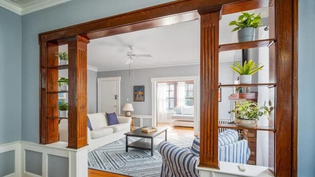 Un salón con un sofá blanco, una mesita de centro y un sillón a rayas. Se observa una estantería de madera con plantas a la derecha.