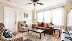 Ein Wohnzimmer mit Deckenventilator, einem braunen Sofa, einem gemusterten Sessel, einem Couchtisch und einem Fernseher.