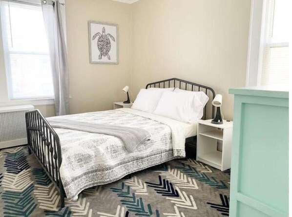 A bedroom with a bed, a nightstand, a lamp, a framed picture on the wall, and a patterned rug.