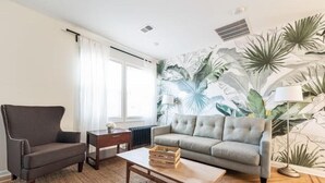 A living room with a grey sofa, a dark armchair, a wooden coffee table, and a large window with white curtains.