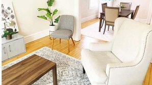 A living room with a grey armchair, a white sofa, a wooden coffee table, a potted plant, and a dining area with a table and chairs. - 2-BDR Oasis Near Beth Isreal Hosp 10 mins to EWR (Newark)