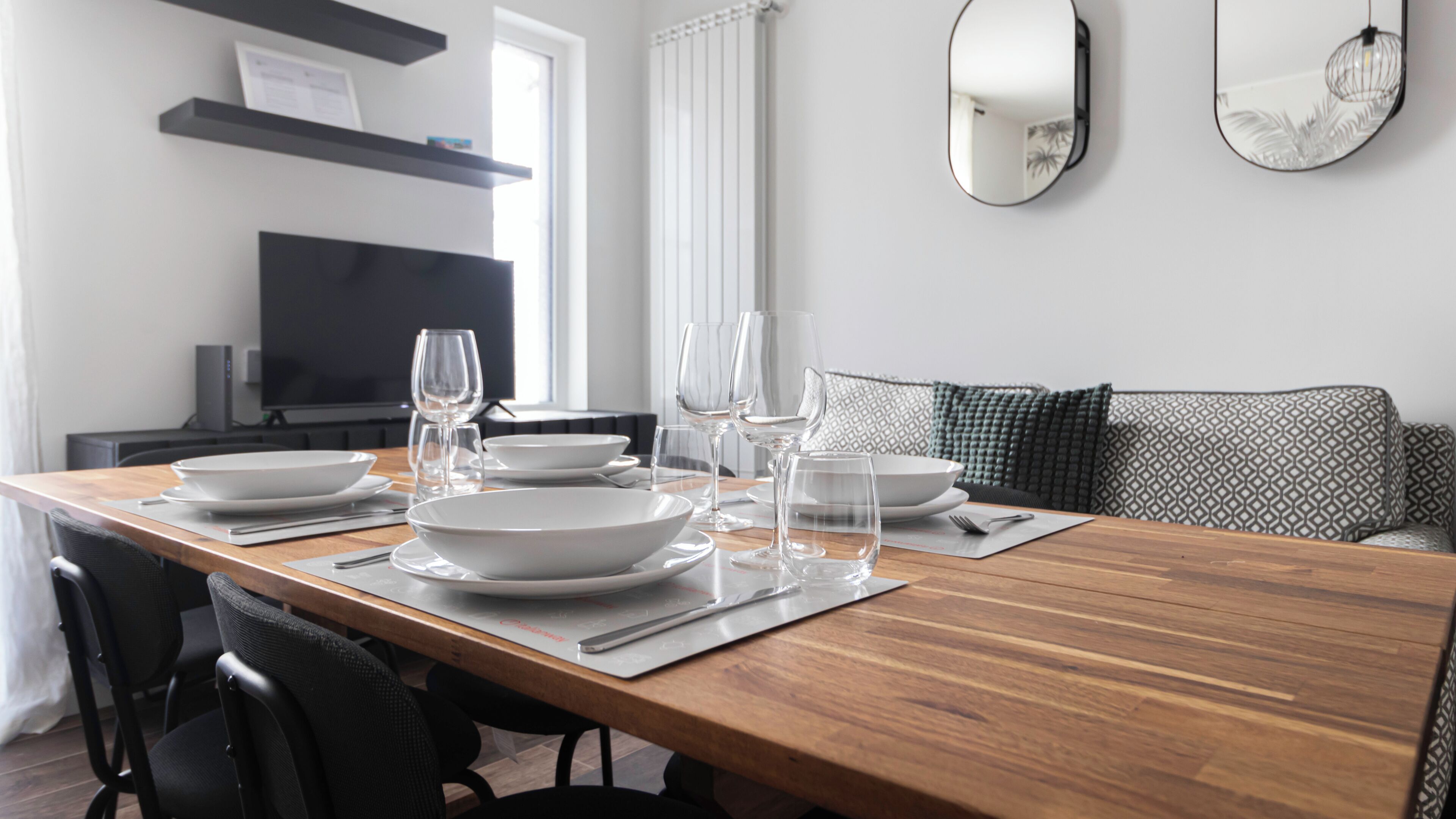A dining table set with white plates, glasses, and cutlery, a black chair, a sofa with patterned cushions, a television, and a mirror on the wall.