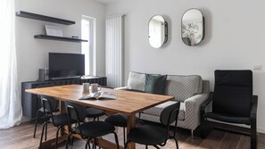 A modern living room with a wooden dining table, black chairs, a sofa, and a black armchair. There is a flat-screen TV on a stand, a radiator, and two wall-mounted mirrors. - Italianway Comfort - Buenos Aires 65 E (Milan)