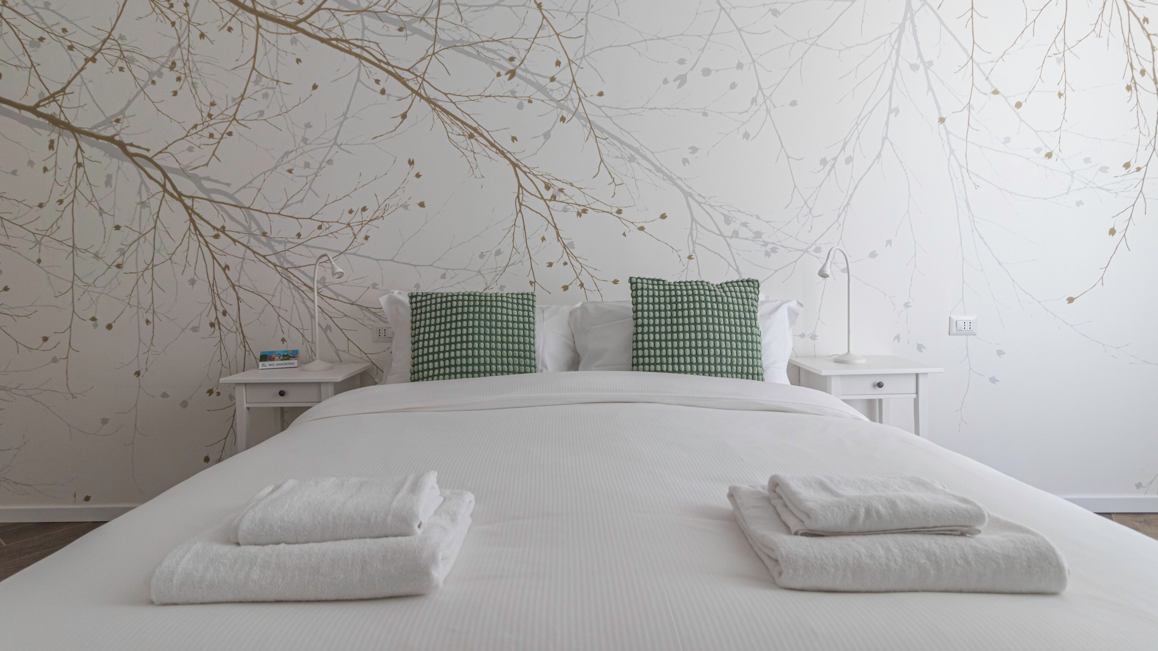 Un letto rifatto con due asciugamani piegati, un comodino bianco con una lampada e una parete decorata con un motivo a rami.