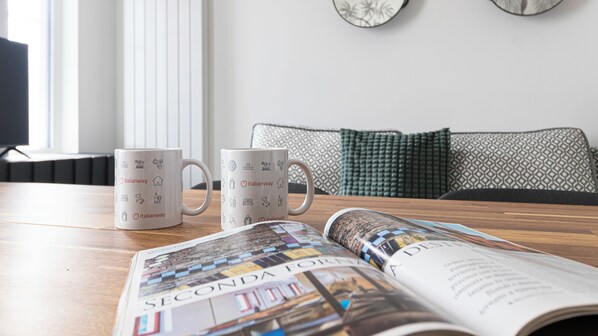 A table with two white mugs featuring travel-related icons, a magazine open to a page with colorful images, and a modern sofa with patterned cushions in the background.