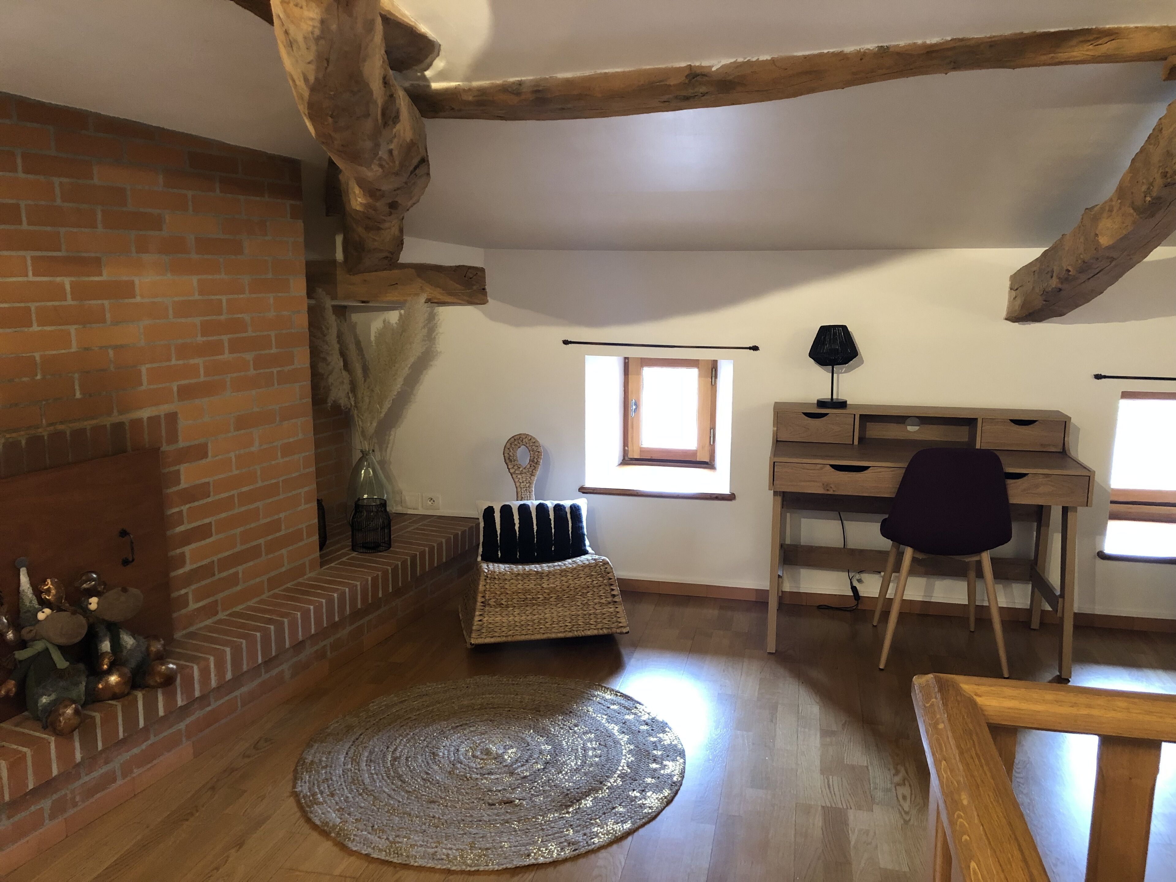 Une pièce chaleureuse avec des poutres apparentes, une cheminée en briques, un bureau avec une chaise et une grande fenêtre.