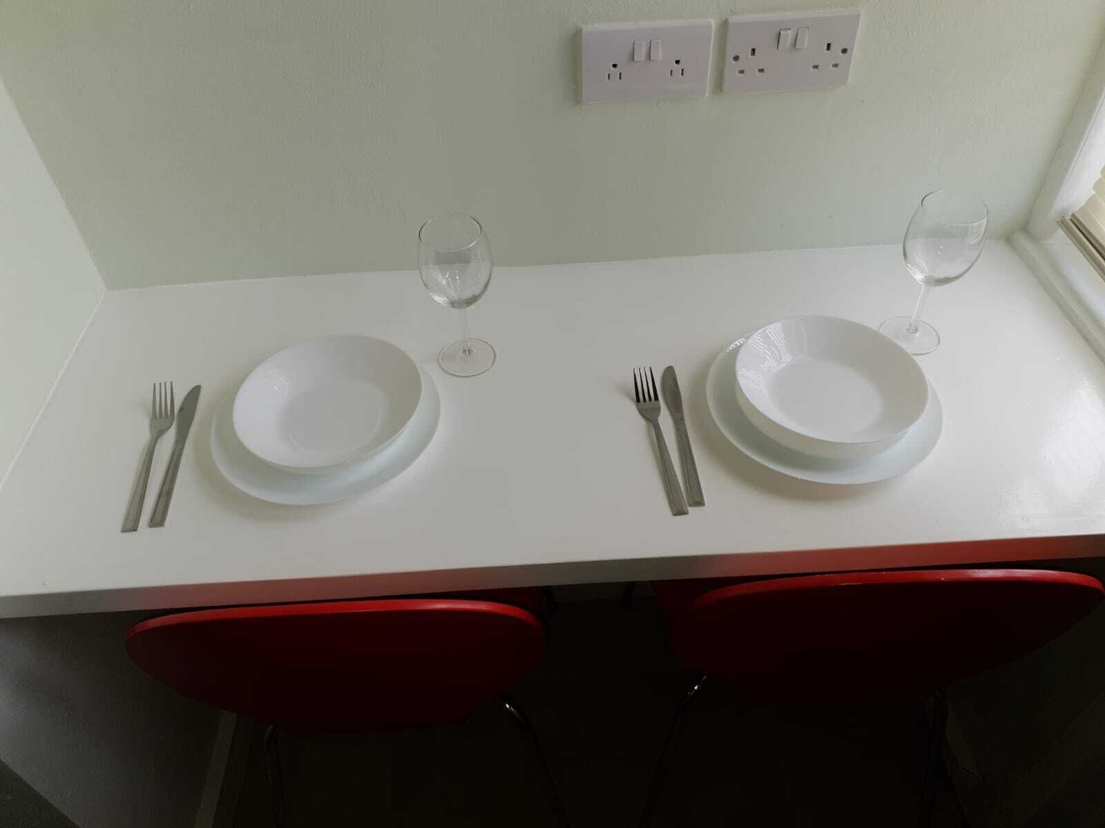 A dining table set with white plates, cutlery, and wine glasses, flanked by red chairs.