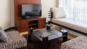 Un salon moderne avec un téléviseur à écran plat posé sur un meuble en bois, deux canapés à motifs, une table basse et une grande fenêtre ornée de rideaux légers.