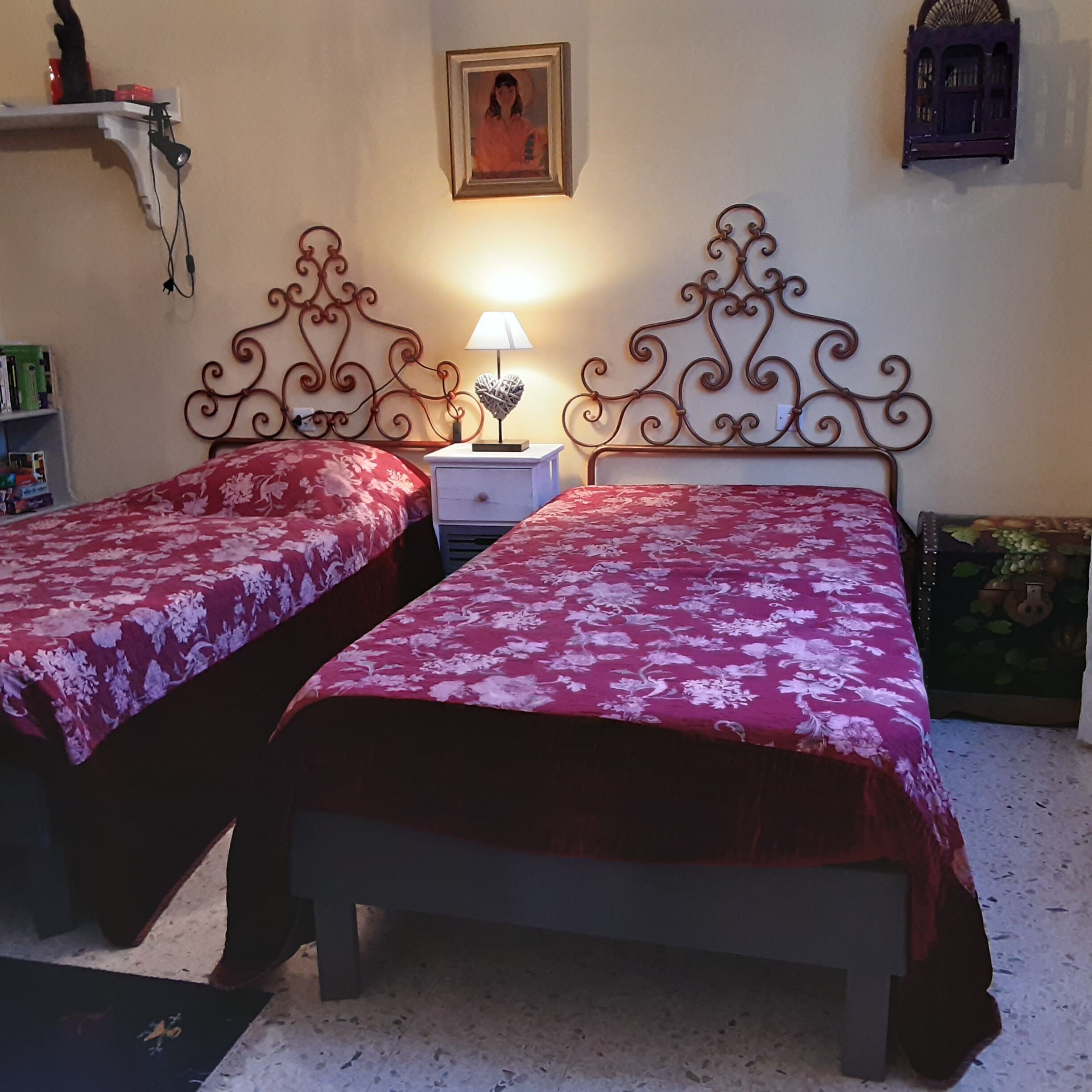 Ein Schlafzimmer mit zwei Betten, einem Nachttisch mit Lampe, einem Regal mit Büchern und einem gerahmten Bild an der Wand.