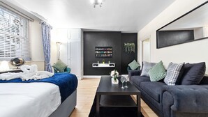 A modern hotel room with a bed, a sofa, a coffee table, a TV, and a wall-mounted shelf with decorative items. - 1 Bed Flat - Sleeps 5 - Pets - Parking - Wifi (London)