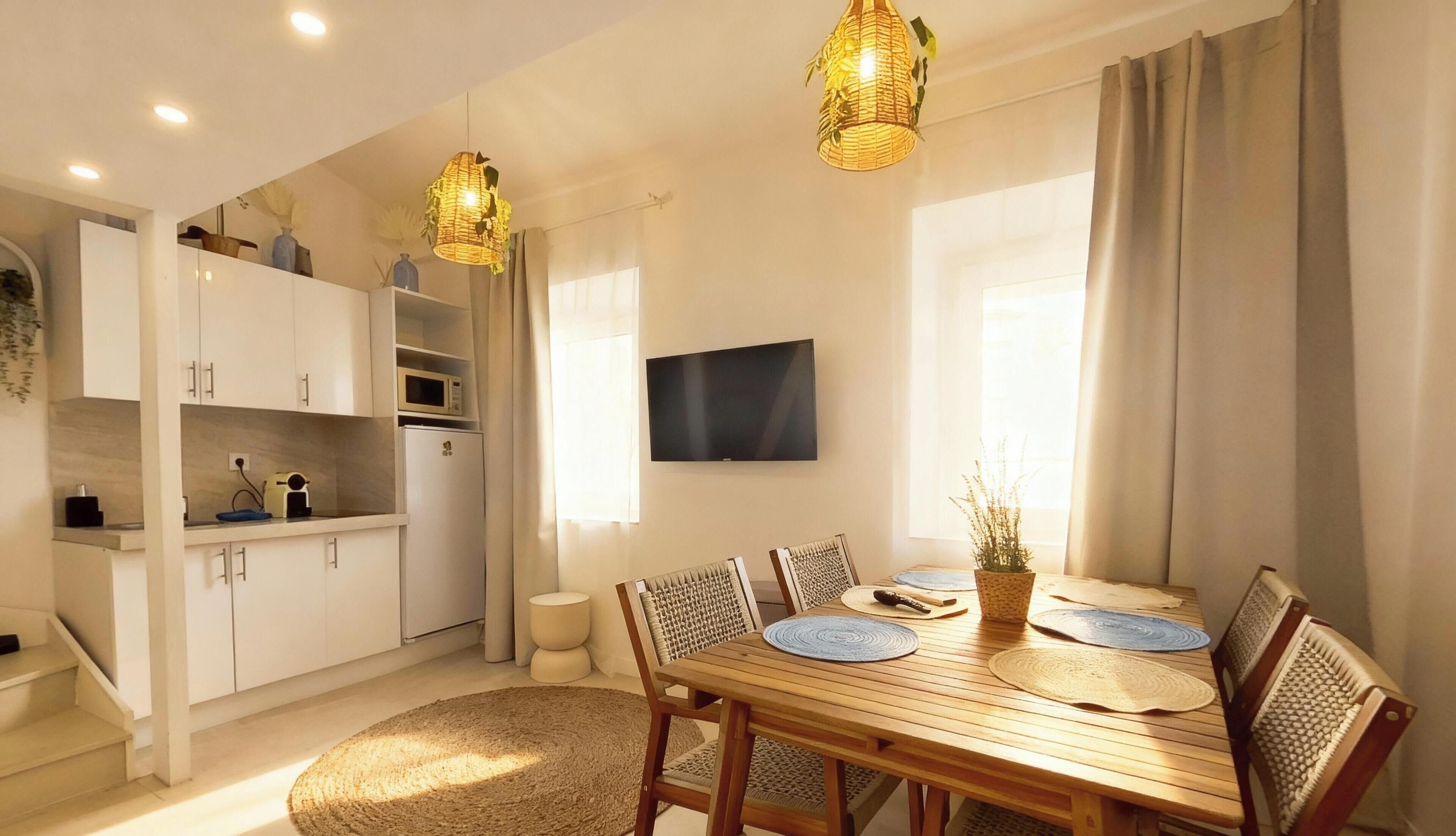 A modern kitchen with white cabinets, a microwave, and a refrigerator. There is a dining table with chairs and a TV mounted on the wall.