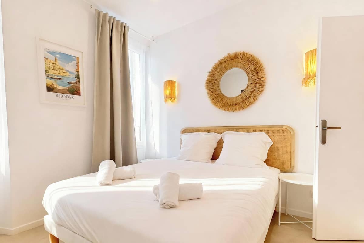 A neatly made bed with white linens and towels, a framed picture of a coastal scene, a round mirror, and a door with a light switch.